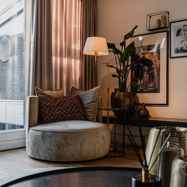 Modernes Wohnzimmer mit rundem Samtsessel, goldfarbener Stehleuchte, Vorhängen und Zimmerpflanze in dekorativem Topf.