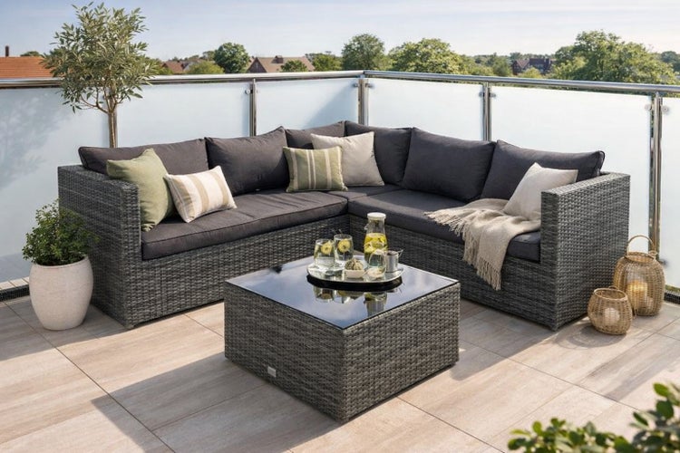 Graue Gartenlounge aus Polyrattan in L-Form mit dunklen Polstern und quadratischem Loungetisch mit Glasplatte auf einer hellen Terrasse.