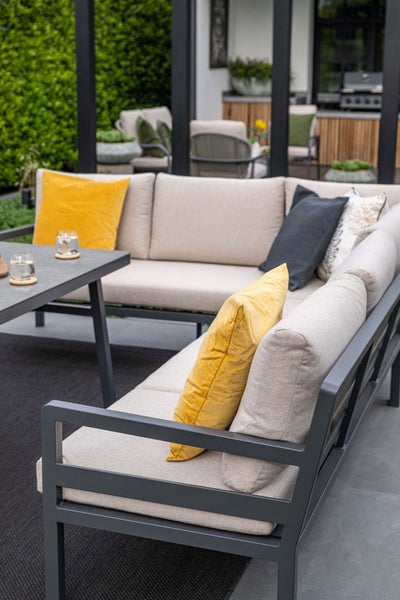 Modernes Garten-Ecksofa mit grauem Aluminiumgestell, beigen Polstern und gelben Zierkissen auf einer Terrasse neben einem passenden Beistelltisch.