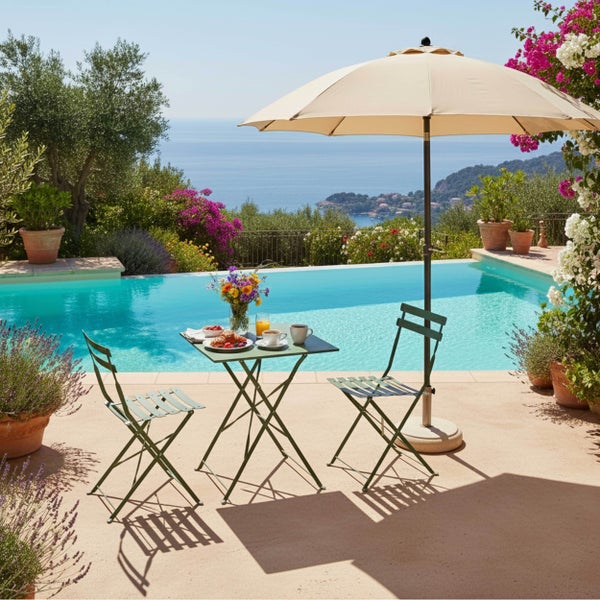 Grüne Bistro-Garnitur aus Metall mit Klapptisch und zwei Stühlen auf sonniger Terrasse am Pool mit beigem Sonnenschirm und Meerblick.