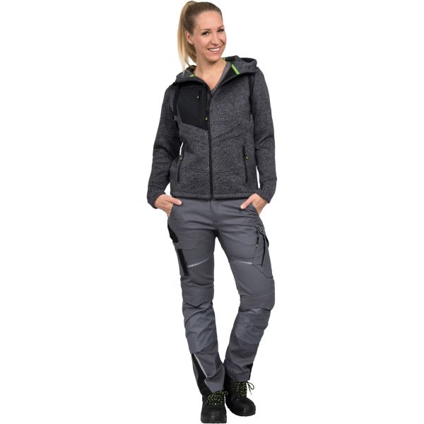 Frau in Arbeitskleidung bestehend aus einer grauen Strickjacke mit Kapuze, einer grauen Arbeitshose mit Taschen und schwarzen Sicherheitsschuhen.
