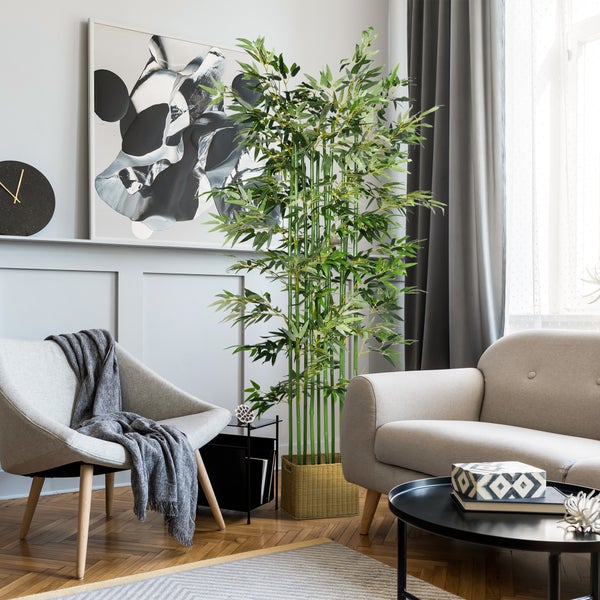 Große künstliche Bambuspflanze im Flechtkorb in einem hellen, modernen Wohnzimmer mit grauem Sessel, Sofa und abstrakter Wandkunst.