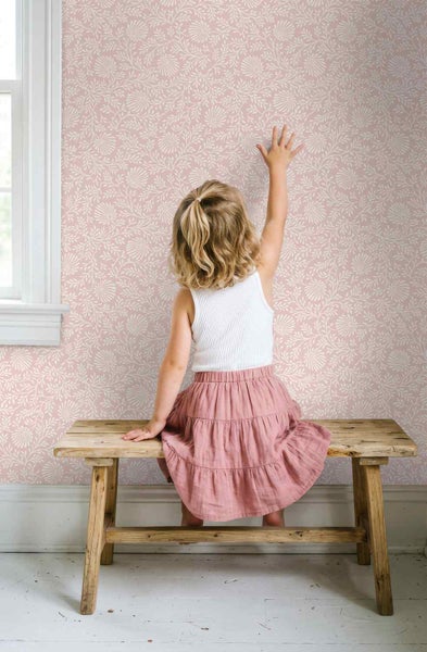 Kind sitzt auf einer Holzbank vor einer Wand mit floraler Vliestapete mit weißem Blumenmuster.