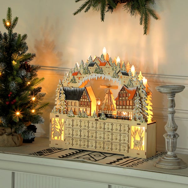 Festlich beleuchteter Adventskalender aus Holz mit Schwibbogen, Winterdorf-Motiv und 24 nummerierten Schubladen auf einer hellen Kommode.