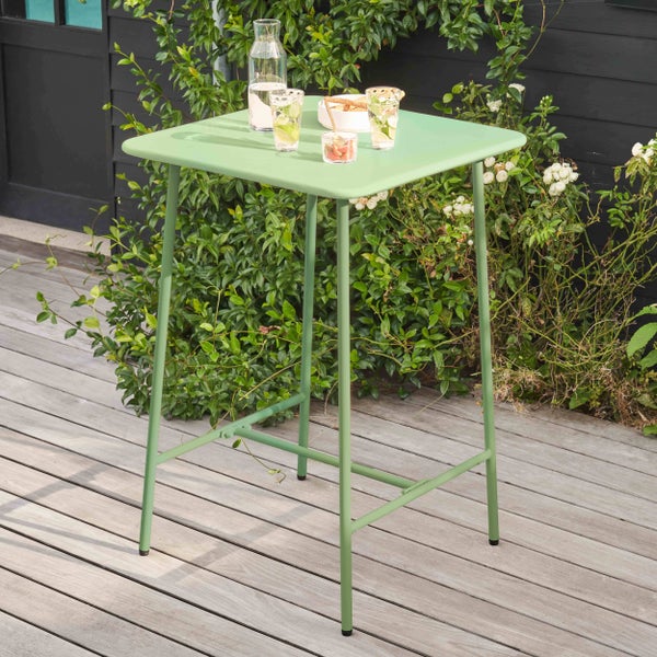 Moderner, quadratischer Stehtisch aus Metall in Grün, stehend auf einer Holzterrasse mit Pflanzen im Hintergrund.