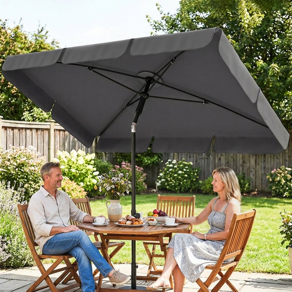 Rechteckiger, dunkelgrauer Sonnenschirm aus Textil und Metall über einem runden Holztisch mit Gartenstühlen auf einer hellen Terrasse im Garten.