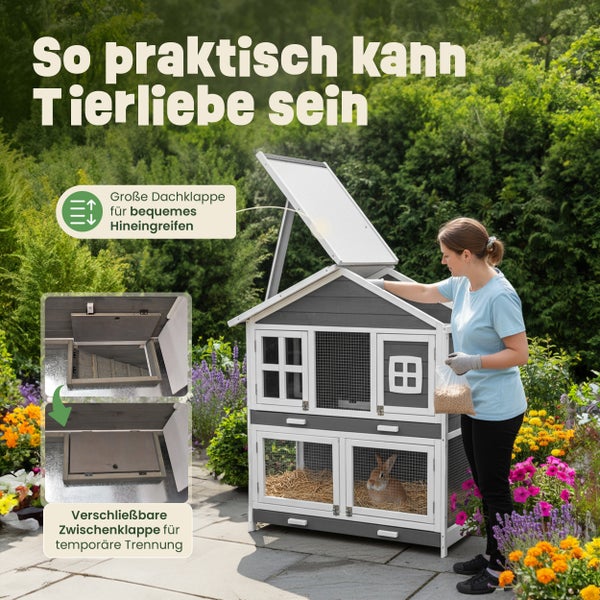 Zweistöckiger grau-weißer Kleintierstall aus Holz im Garten mit geöffneter Dachklappe zur Fütterung und verschließbarer Zwischenklappe zur Trennung.
