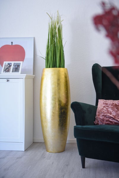 Hohe, goldfarbene Bodenvase mit künstlichem Gras in einem modern eingerichteten Wohnzimmer neben einem dunklen Sessel und einem weißen Schrank.