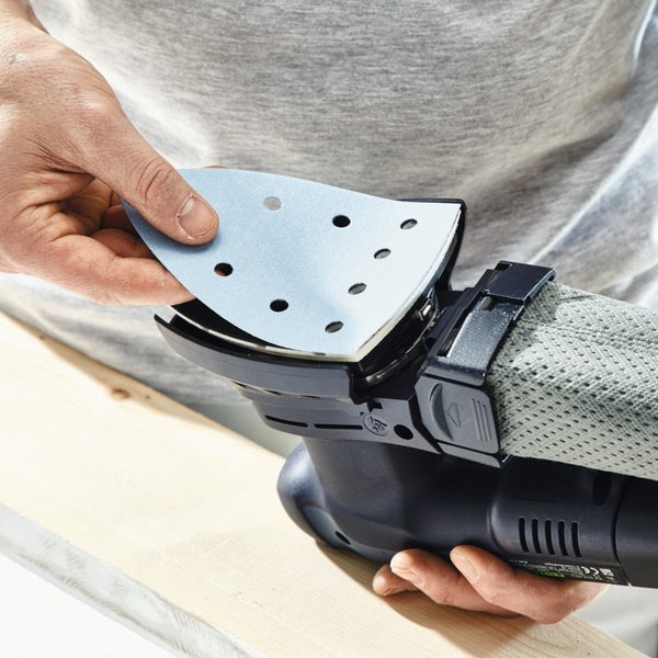 Hand befestigt ein dreieckiges Schleifblatt an einem Deltaschleifer mit Staubbeutel über einem Holzbrett.
