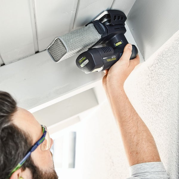 Ein Mann schleift mit einem akkubetriebenen Festool Deltaschleifer eine Holzbalkendecke über Kopf, inklusive Staubfangbeutel und Schutzbrille.