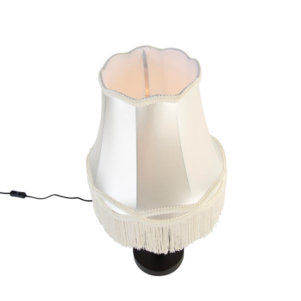 Tischleuchte mit beigefarbenem Stoffschirm, gewelltem Rand, Fransen, schwarzem Standfuß und Kabel mit Schnurschalter.