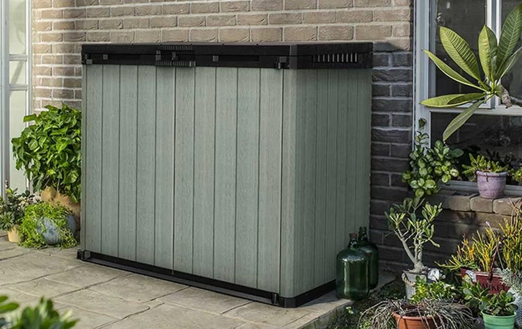 Gartenbox mit grau-grünen Paneelen in Holzoptik und schwarzem Deckel auf einer Terrasse vor einer Ziegelwand.