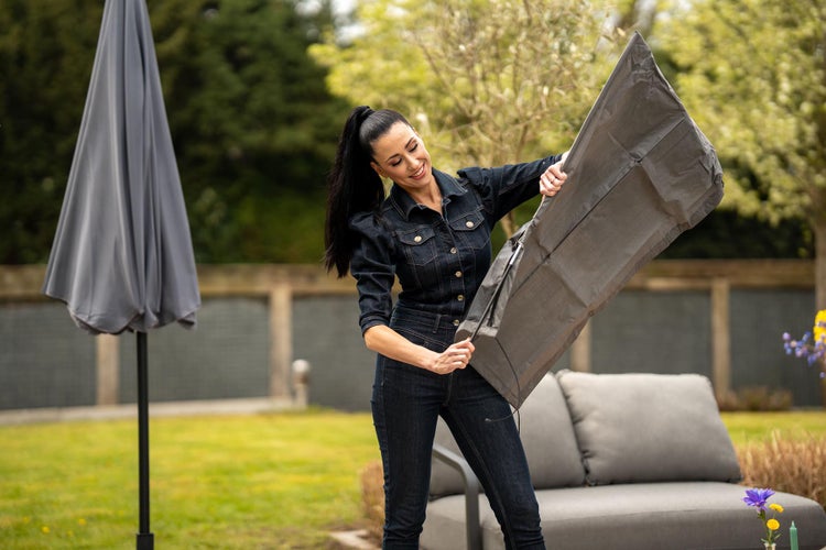 Szene im Garten mit einer Frau, die eine Schutzhülle für einen Sonnenschirm hält, mit einem Sonnenschirm und einer grauen Lounge im Hintergrund