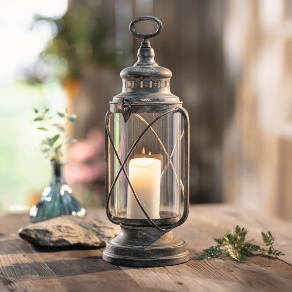 Dekorative Laterne aus Metall im Antiklook mit brennender Stumpenkerze in einem Glaszylinder auf einem Holztisch.
