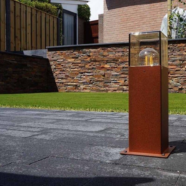 Moderne Sockelleuchte in Rostoptik mit Glaseinsatz und sichtbarem Leuchtmittel auf einer Terrasse im Garten mit Steinwand und Rasen.