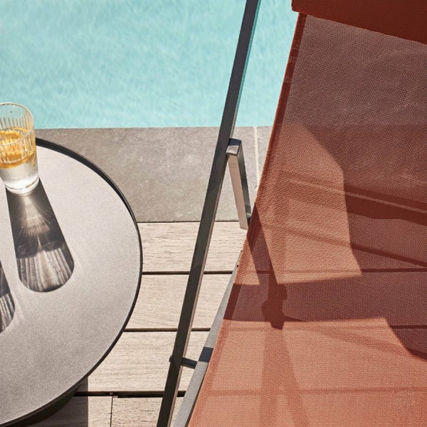 Sonnenliege mit terracottafarbenem Netzgewebe und dunklem Metallgestell neben einem runden Beistelltisch auf einer Holzterrasse am Pool.