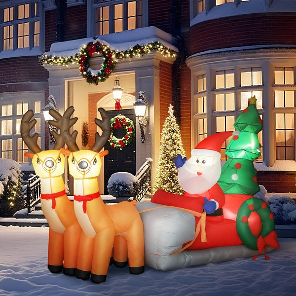 Aufblasbare Weihnachtsdekoration mit Weihnachtsmann im Schlitten, zwei Rentieren und einem Tannenbaum, beleuchtet vor einem Haus im Schnee.