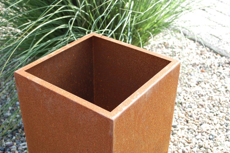 Quadratischer Pflanzkübel aus Cortenstahl in Rostoptik in einem Garten mit Kies und Ziergras.