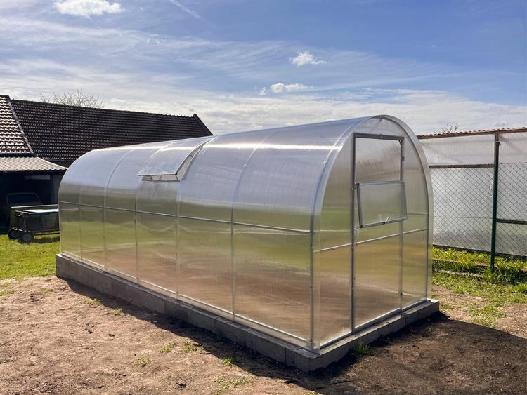 Langes Tunnelgewächshaus mit transparenten Kunststoffplatten und Metallrahmen, auf einem Fundament aus Betonsteinen im Garten platziert.
