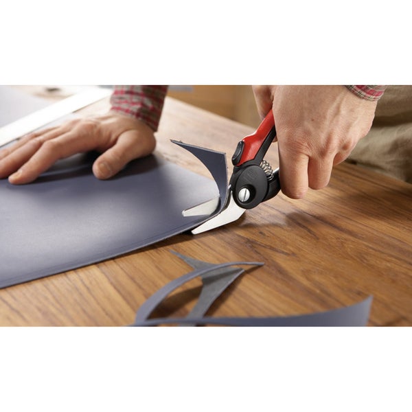 Eine Person schneidet mit einer Blechschere ein Stück Material auf einem Holztisch.