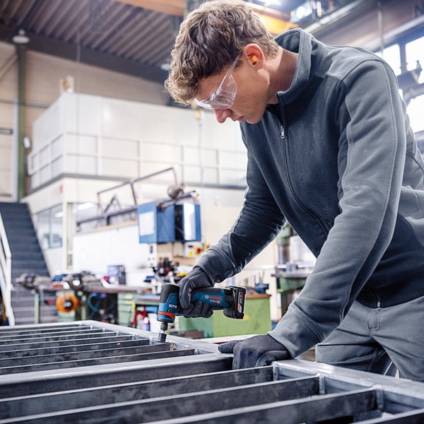 Mann arbeitet mit einem Akku-Bohrschrauber an einem Metallrahmen, getragen sind Schutzbrille und Handschuhe.