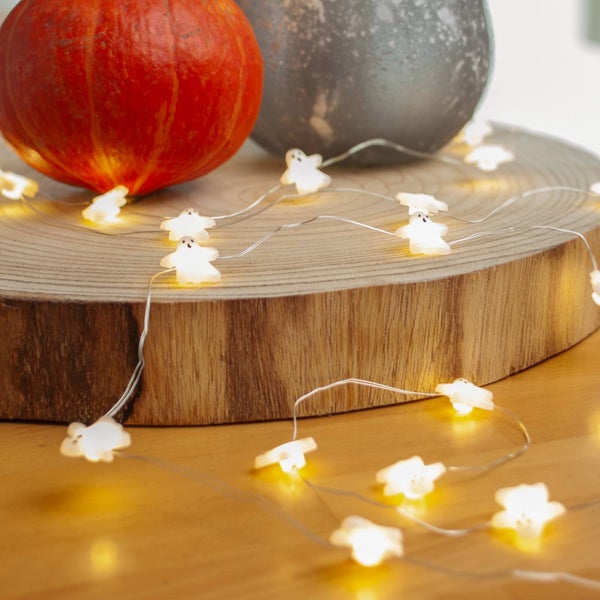 LED-Lichterkette mit kleinen leuchtenden Gespenstern auf einer Baumscheibe aus Holz neben Kürbissen.
