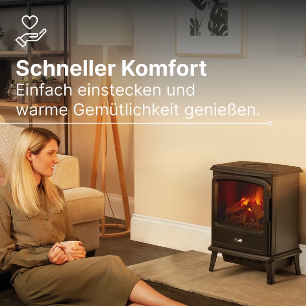 Eine Frau sitzt mit einer Tasse vor einem schwarzen elektrischen Kaminofen mit Flammeneffekt in einem hellen Wohnzimmer.