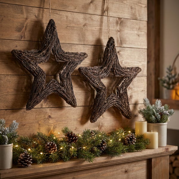 Zwei hängende Weihnachtssterne aus geflochtenen Zweigen vor einer Holzwand mit Tannengirlande und Kerzen als festliche Weihnachtsdekoration.