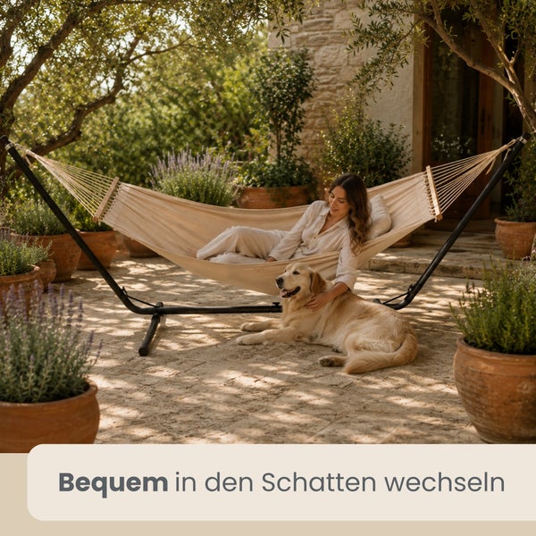 Frau und Hund auf Steinterrasse mit Tuchhängematte auf schwarzem Metallgestell, umgeben von Tontöpfen mit Lavendel unter schattigen Bäumen.