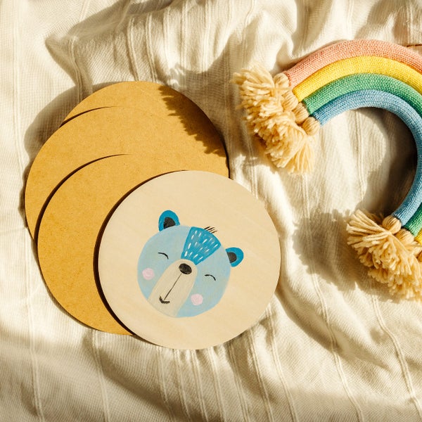 Runde Holzscheiben mit Bären-Illustration und ein dekorativer Regenbogen auf einer strukturierten Stoffunterlage.