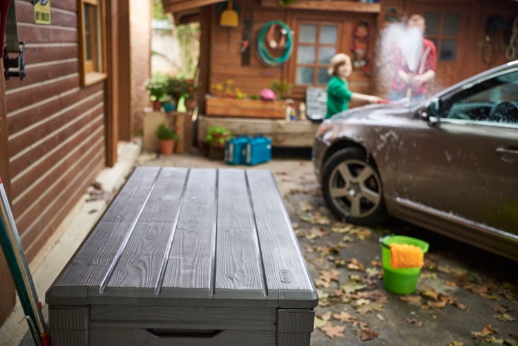 Eine graue Aufbewahrungsbox im Freien, mit Personen beim Autowaschen im Hintergrund.