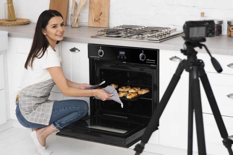 Frau kniet in einer modernen Küche vor einem eingebauten Backofen und schiebt ein Backblech mit Gebäck hinein während sie vor einer Kamera backt.
