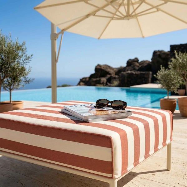Gestreifter Outdoor-Hocker an einem Pool unter einem grossen Sonnenschirm in einer hellen Gartenanlage mit Olivenbäumen.