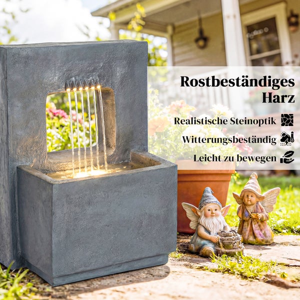 Moderner Gartenbrunnen in grauer Steinoptik aus Kunstharz mit LED-Beleuchtung und fließendem Wasser neben Gartenfiguren in einer Gartenanlage.