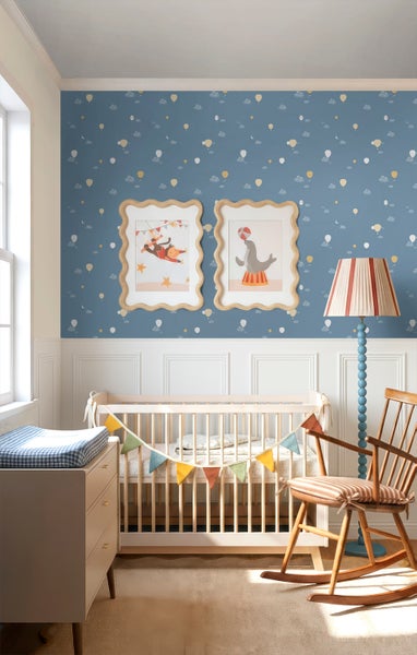 Hell gestaltetes Babyzimmer mit blauer Vliestapete mit Heißluftballon-Muster, weißer Wandvertäfelung, Holz-Gitterbett und Schaukelstuhl.