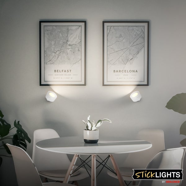 Zwei kleine weiße Wandleuchten beleuchten gerahmte Bilder in einem modern eingerichteten Esszimmer. StickLights by Briloner Logo.