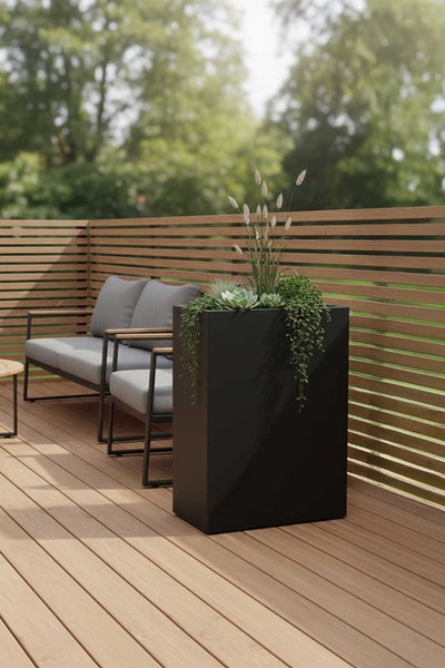 Moderner, rechteckiger Pflanzkübel in Schwarz auf einer Holzterrasse mit verschiedenen Pflanzen, grauen Gartenmöbeln und einem Holzlamellenzaun.