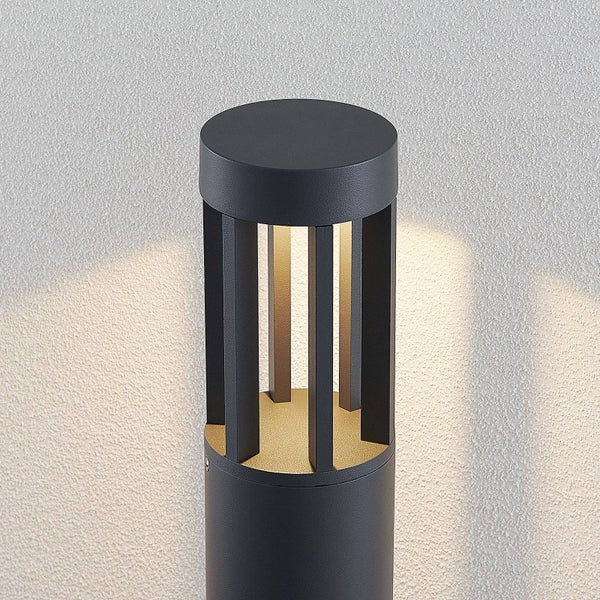 Moderne Außenleuchte in Anthrazit, zylindrische Form mit vertikalen Lamellen und warmweißem Lichtkegel an einer Wand.