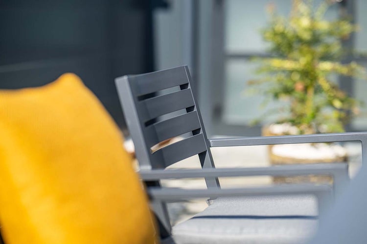 Nahaufnahme eines modernen anthrazitfarbenen Gartenstuhls mit Lattenrückenlehne, grauem Sitzkissen und gelbem Dekokissen im Außenbereich.