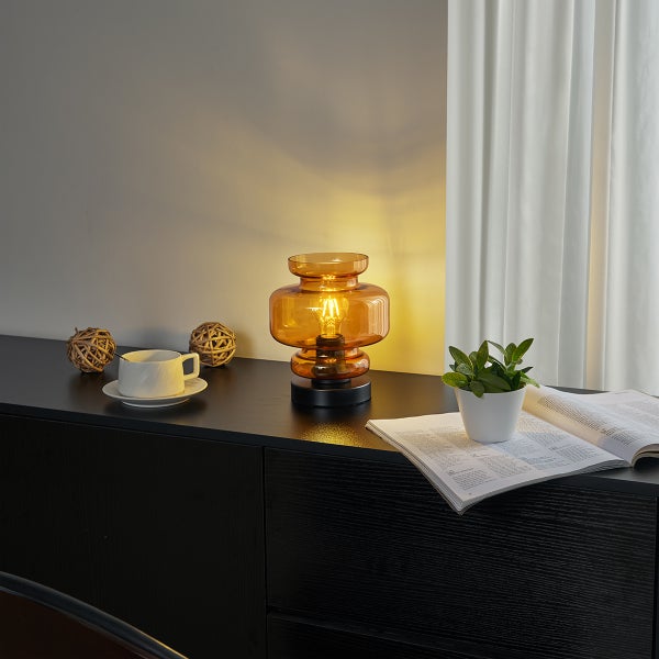 Dekorative Tischleuchte aus Glas auf einem Sideboard mit Dekorationen und Zimmerpflanze.