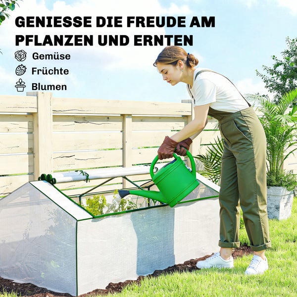 Eine Frau gießt Gemüse, Früchte und Blumen in einem kleinen Foliengewächshaus im Garten mit einer Gießkanne.