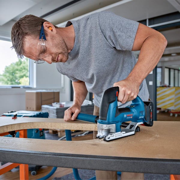 In einer hellen Werkstatt schneidet ein Handwerker mit Schutzbrille eine Aussparung in eine Holzplatte mit einer Bosch Professional Akku-Stichsäge inklusive Staubabsaugung.
