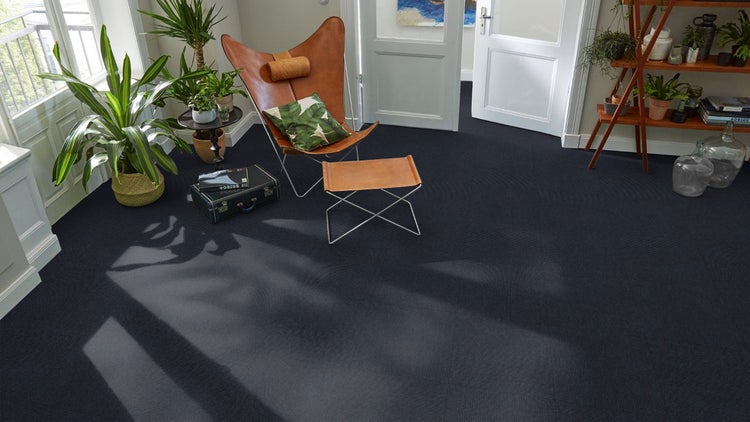 Modernes Wohnzimmer mit dunkelblauen Teppichfliesen, Leder-Schmetterlingsstuhl, Fußhocker, Zimmerpflanzen und natürlichem Licht.