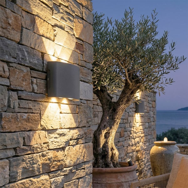 Moderne Außenwandleuchte an einer Steinmauer mit Lichtkegeln nach oben und unten neben einem Olivenbaum im Topf bei Dämmerung.
