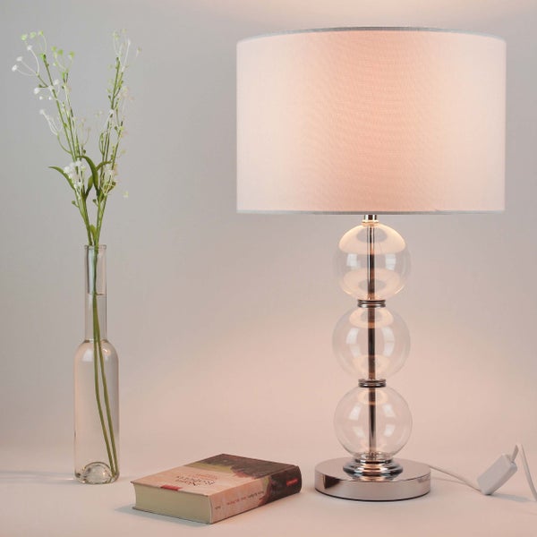 Dekorative Tischleuchte mit Stoffschirm und Glasfuß, neben einem Buch und einer Vase mit Blumen.