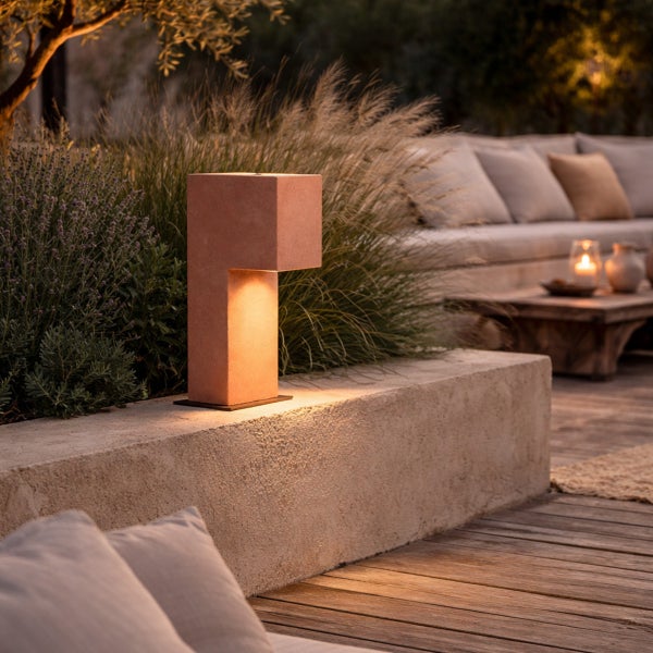 Moderne Sockelleuchte aus Beton in L-Form, platziert auf einer Gartenmauer, abends beleuchtet mit gemütlichem Lichtschein auf der Holzterrasse.