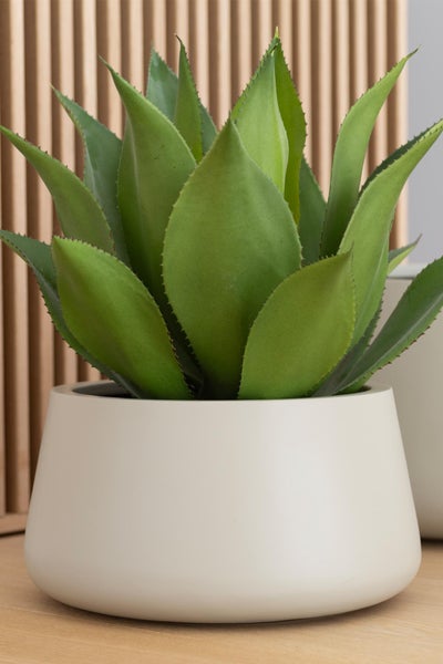 Künstliche Agave im cremefarbenen Übertopf vor einer Holzlamellenwand.