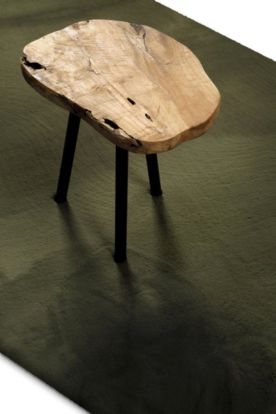 Beistelltisch aus einer massiven Baumscheibe mit drei schwarzen Beinen auf einem dunkelgrünen Hochflorteppich.