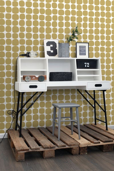 Weißer Schreibtisch mit Regalen, schwarzem Metallgestell und grauem Hocker auf einer Holzpalette vor einer gemusterten Tapete.