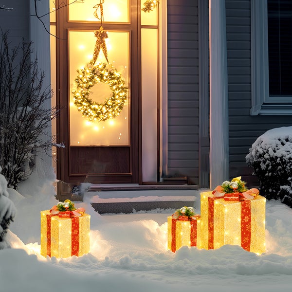 Beleuchtete LED-Geschenkboxen im Schnee vor einer Haustür mit leuchtendem Weihnachtskranz, festliche Außendekoration.